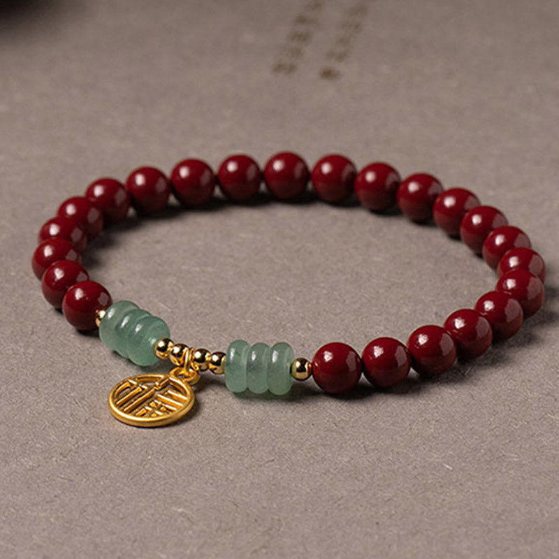 Armband Grüner Aventurin Zinnober auf grauem Untergrund liegend, rote Perlen mit grünen Steinen und goldenem Anhänger.