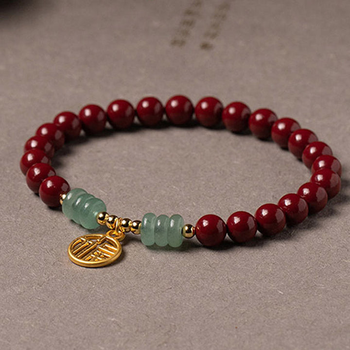 Armband Grüner Aventurin Zinnober auf grauem Untergrund liegend, rote Perlen mit grünen Steinen und goldenem Anhänger.