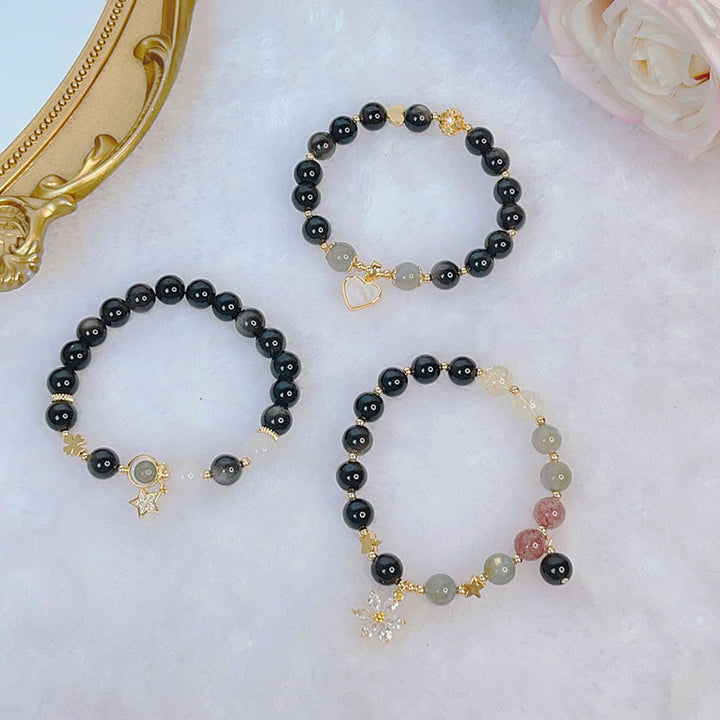 Drei Obsidian Armbänder mit goldenen Charms auf weißem Hintergrund.