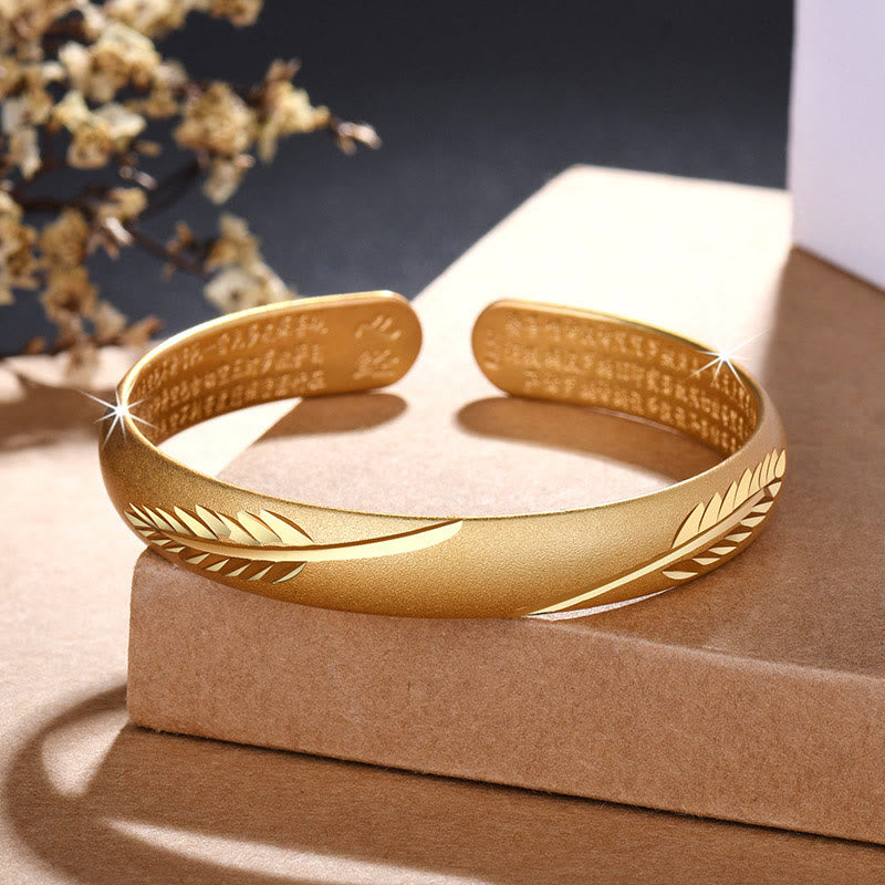 Goldenes Armband mit Feder-Design auf beigem Stein displayiert elegant.