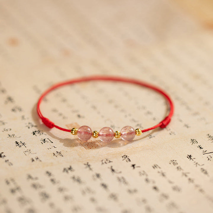 Armband Erdbeerquarz auf Buch mit roten Fäden goldenen Perlen Glück Lebensfreude