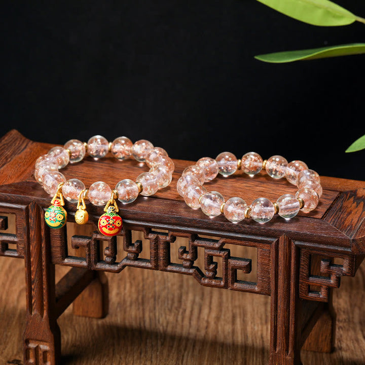 Rosenquarz Armband mit vergoldeten Glücksanhängern auf Holzbox.