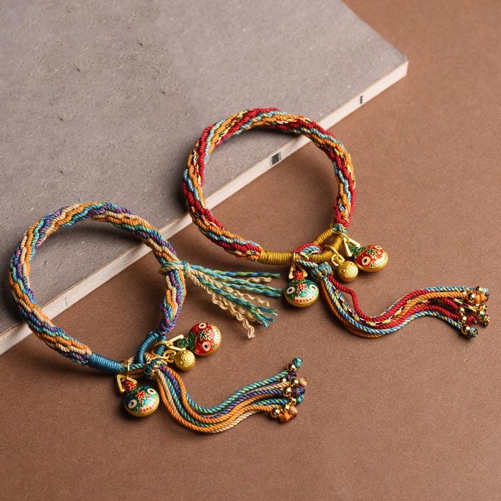 Bunte Seidenfaden Armbänder mit goldenen Glöckchen Anhängern auf beigem Hintergrund.