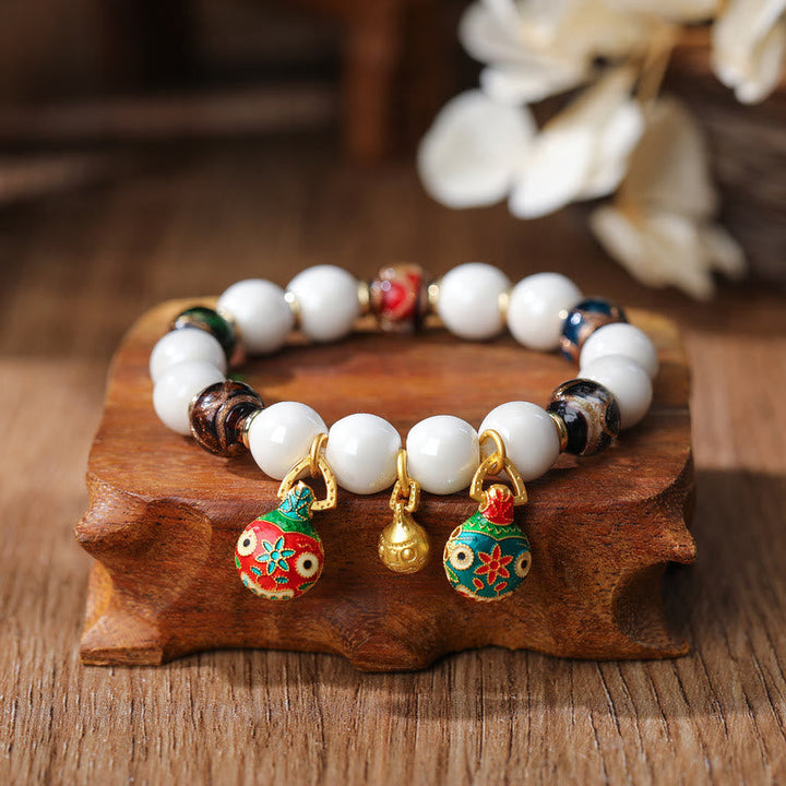 Weißes Perlenarmband mit vergoldeten Glücksbringern auf Holzständer.