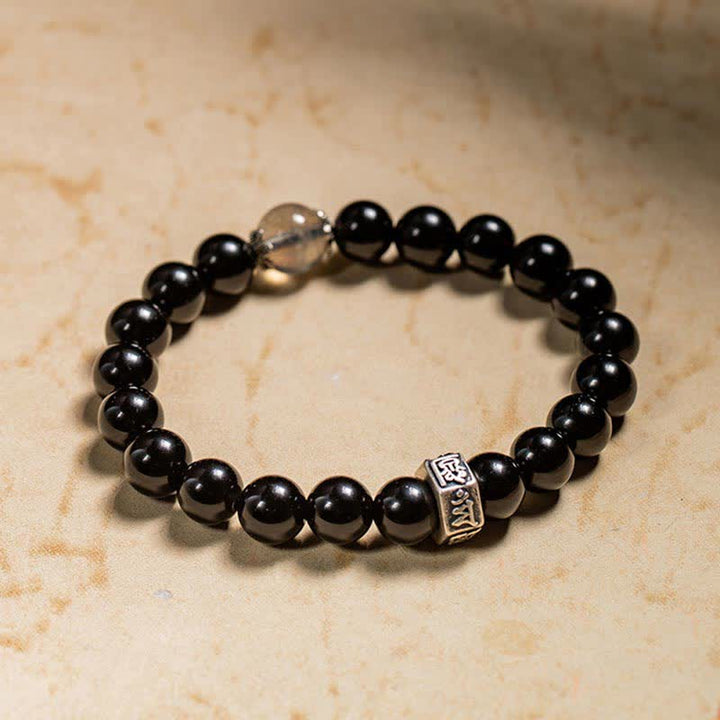Schwarzes Obsidian Armband mit Silber Charm auf beigem Hintergrund elegant.